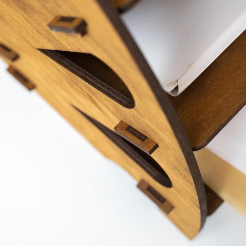 K&ouml;nnte beinhalten: Nahaufnahme eines Schreibtisch-Organizers aus Holz mit hellbraunem Finish. Der Organizer hat ein einzigartiges Design mit ausgeschnittenen Formen und dunkelbraunen Akzenten. Die Konstruktion umfasst kleine Holzd&uuml;bel zur Montage. Ein wei&szlig;es Objekt ist teilweise sichtbar.
