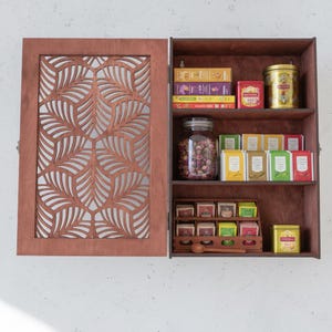 May include: A wooden tea cabinet with a decorative leaf-patterned door. The open cabinet displays various tea boxes, tins, and a jar of dried rosebuds. The cabinet is made of brown wood and is mounted on a white wall.