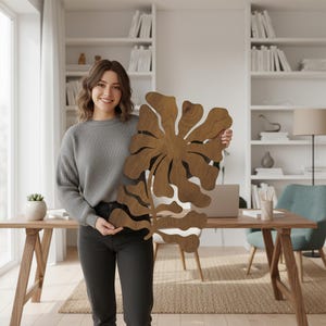 Peut inclure: Une grande &oelig;uvre d'art murale florale en bois avec une finition en grain de bois naturel. Le design pr&eacute;sente une forme stylis&eacute;e de fleur et de feuille. L'&oelig;uvre est tenue par une personne en pull gris et jean noir.