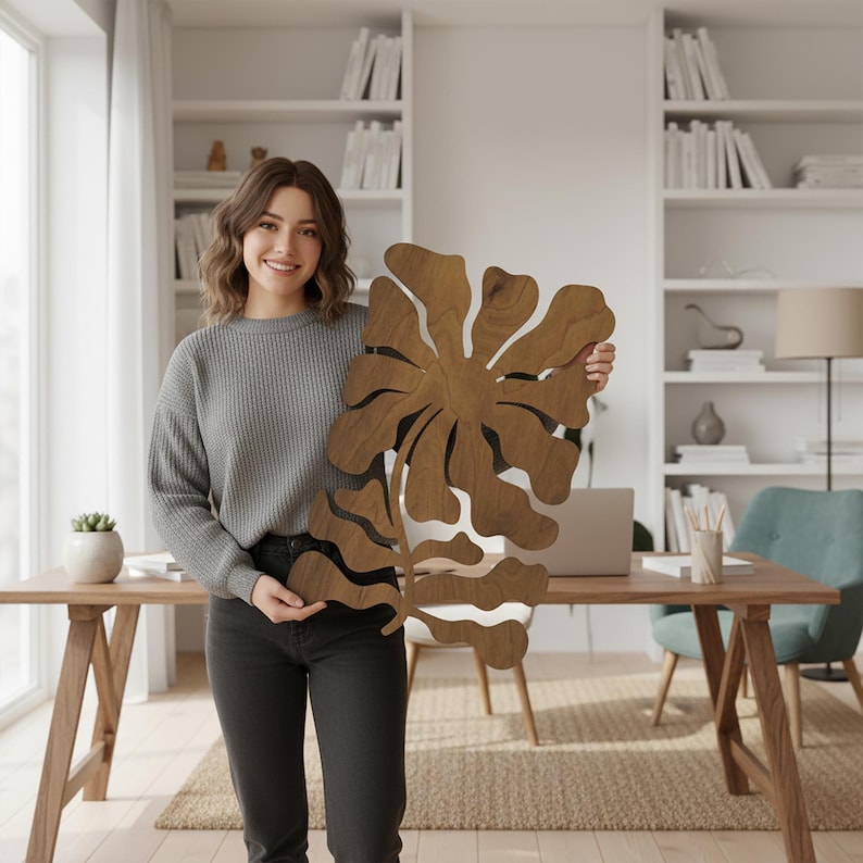 Peut inclure: Une grande &oelig;uvre d'art murale florale en bois avec une finition en grain de bois naturel. Le design pr&eacute;sente une forme stylis&eacute;e de fleur et de feuille. L'&oelig;uvre est tenue par une personne en pull gris et jean noir.