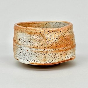 May include: A handmade ceramic bowl with a unique, textured surface. The bowl features a square shape with rounded corners and a wide opening. The exterior is a blend of orange, brown, and white, with a crackled pattern. The bowl sits on a small, dark brown base.