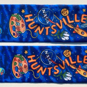 May include: A pair of blue socks with a tie-dye pattern. The socks feature a variety of colorful designs, including a rocket ship, a paint palette, a flower, and the words "Huntsville" and "NASA".