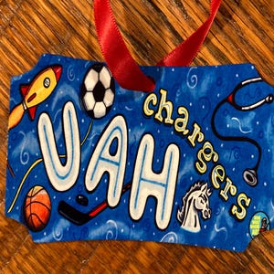 May include: A blue and white fabric ornament with the name "VAH" in large white letters with a blue outline. The ornament also features a soccer ball, basketball, hockey stick, rocket, stethoscope, and a white horse. The text "chargers" is written in yellow letters on the ornament.