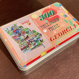 May include: A metal tin box with a colorful illustration of the state of Georgia. The box contains a 300 piece jigsaw puzzle. The text on the box reads "The Real What & Where of Georgia" and "300 Piece Jigsaw Puzzle Georgia".