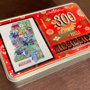 May include: A red, white, and blue tin box with a 300 piece jigsaw puzzle of Mississippi. The box has a floral design and the text "300 Piece Jigsaw Puzzle - Mississippi".