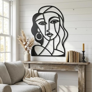 May include: A black metal wall art piece featuring a continuous line drawing of a stylized human face with long flowing hair and a circular earring. The design is composed of bold, dark lines that form the facial features and hair contours.