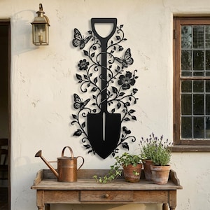Può includere: Un'opera d'arte da parete in metallo nero a forma di vanga, caratterizzata da intricati intagli di viticci, foglie, fiori e tre farfalle. Il design è simmetrico, con il manico della vanga in alto e la lama nella parte inferiore.