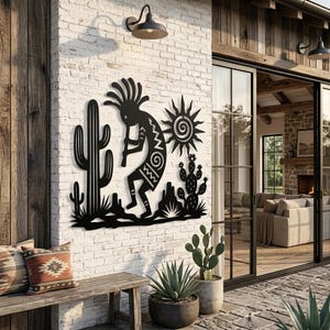 Op de afbeelding: Een zwart metalen wanddecoratie met het silhouet van een Kokopelli-figuur die fluit speelt, geplaatst tussen een hoge saguaro-cactus en een kleinere schijfcactus, met een gestileerd zonontwerp boven het woestijnlandschap.