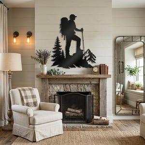 May include: A black metal wall art piece featuring a silhouette of a hiker with a backpack and trekking pole standing on a rocky mountain peak next to pine trees. The design is mounted on a light-colored horizontal wood plank wall above a fireplace.