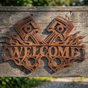 Peut inclure: Une enseigne murale en métal rouillé portant le mot WELCOME en lettres capitales, flanqué de deux pistons de moteur et de motifs de flammes stylisés. L'enseigne a une finition brune oxydée et patinée sur une surface en planches de bois.