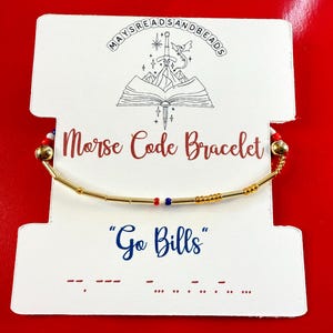 May include: A gold-colored Morse code bracelet with red and blue beads, displayed on a white card with the text "Morse Code Bracelet" and "Go Bills". The card has a logo at the top and is set against a red background.