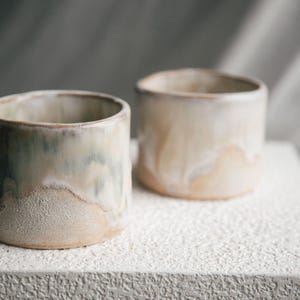 Puede incluir: Dos tazas de cerámica hechas a mano con forma cilíndrica. Las tazas tienen una paleta de colores neutros, con una mezcla de tonos beige, crema y verde claro. La superficie texturizada añade un toque rústico y artesanal a la cerámica.