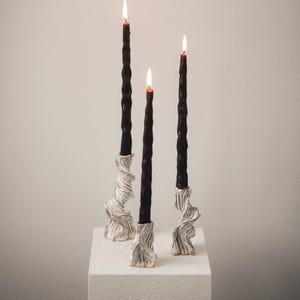 Könnte beinhalten: Drei hohe, schlanke schwarze Stabkerzen mit gedrehter Spiralstruktur, jede mit einer kleinen Flamme brennend. Die Kerzen stecken in drei separaten, weißen Kerzenhaltern aus Keramik, die eine markante, gerüschte und organische skulpturale Form aufweisen.