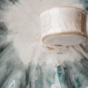 K&ouml;nnte beinhalten: Ein Keramikdeckel mit gl&auml;nzender wei&szlig;er Glasur und einem texturierten, handgeformten Knaufgriff. Die Oberfl&auml;che zeigt ein radiales Muster aus verwaschenen blauen und grauen aquarellartigen Streifen, die von der Mitte zu den R&auml;ndern verlaufen.