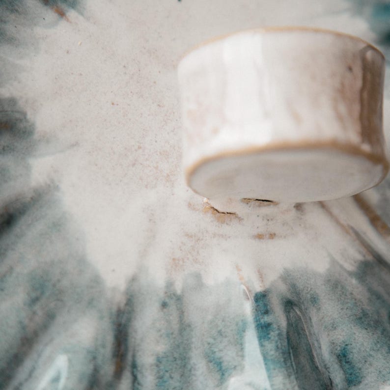K&ouml;nnte beinhalten: Ein Keramikdeckel mit gl&auml;nzender wei&szlig;er Glasur und einem texturierten, handgeformten Knaufgriff. Die Oberfl&auml;che zeigt ein radiales Muster aus verwaschenen blauen und grauen aquarellartigen Streifen, die von der Mitte zu den R&auml;ndern verlaufen.