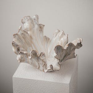 Op de afbeelding: Een keramische kom met een glanzende, gebroken witte afwerking en bruinachtige randen. De kom heeft een sterk onregelmatige, golvende en organische vorm die doet denken aan een groot, open blad of een schelp met diepe, vloeiende plooien.