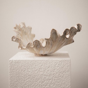 May include: A ceramic bowl with a wide, ruffled, and undulating rim. The surface features a glossy, off-white glaze with subtle brown and grey tonal variations that highlight the organic, flowing curves of the vessel's flared, shell-like structure.