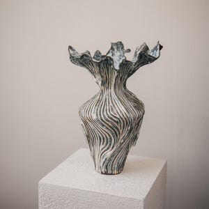 Peut inclure: Un vase en céramique avec un bord évasé et ondulé ainsi qu'un corps effilé. La surface présente un motif texturé à côtes verticales dans des tons blanc cassé, gris et anthracite foncé, créant un effet visuel fluide et contrasté.