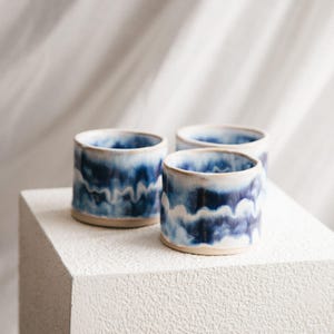 Op de afbeelding: Drie handgemaakte keramische bekers met een cilindrische vorm. De bekers hebben een blauw-wit golfpatroon aan de buiten- en binnenkant, met een natuurlijke, ongeglazuurde basis. De bekers worden gepresenteerd op een wit, getextureerd oppervlak.