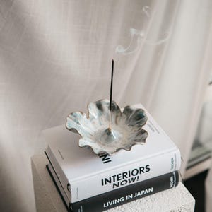 May include: A floral-shaped ceramic incense holder in shades of blue and white, with a gold edge. An incense stick is lit, producing smoke. The holder sits on a stack of books, one titled "INTERIORS NOW!"