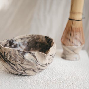 Op de afbeelding: Een keramische matcha kom met een marmerpatroon in beige en donkerbruin. Een bamboe matcha klopper staat op de achtergrond. De kom heeft een onregelmatige vorm, ideaal voor theeceremonies.