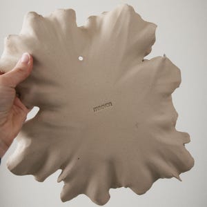 K&ouml;nnte beinhalten: Eine beige Keramik-Wandplatte in Form eines gro&szlig;en, unregelm&auml;&szlig;igen Blattes mit welligen, strukturierten R&auml;ndern. Die Oberfl&auml;che ist matt und weist ein kleines kreisf&ouml;rmiges Loch zum Aufh&auml;ngen oben mittig sowie das Wort maevon auf.