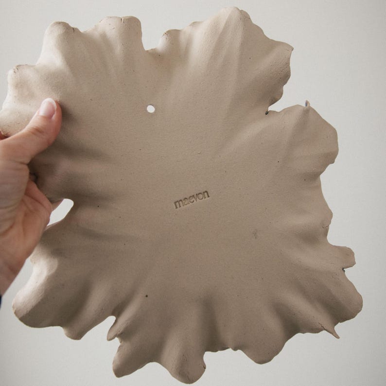 K&ouml;nnte beinhalten: Eine beige Keramik-Wandplatte in Form eines gro&szlig;en, unregelm&auml;&szlig;igen Blattes mit welligen, strukturierten R&auml;ndern. Die Oberfl&auml;che ist matt und weist ein kleines kreisf&ouml;rmiges Loch zum Aufh&auml;ngen oben mittig sowie das Wort maevon auf.