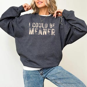 May include: Dark heather grey sweatshirt with the text "I COULD BE MEANER" in a distressed font. The sweatshirt is a relaxed fit and has long sleeves. The bottom of the image shows the words "dark heather grey".