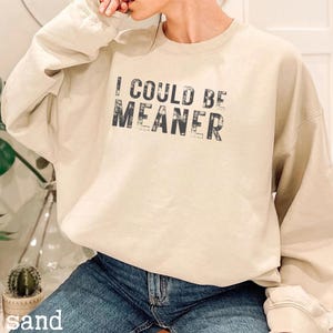 May include: Sand-colored sweatshirt with the text "I COULD BE MEANER" in distressed black lettering. The long-sleeved top is made of a soft material and is paired with blue jeans.