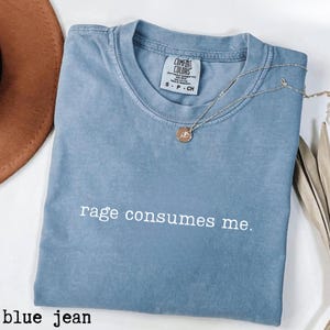 May include: Blue jean colored t-shirt with the phrase "rage consumes me" in white lettering. A silver necklace with a small pendant rests on the shirt. The shirt has a Comfort Colors tag.