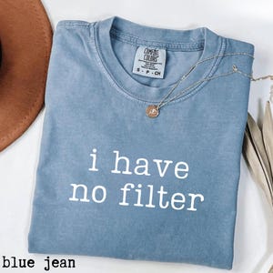 May include: A blue jean colored t-shirt with the phrase "i have no filter" printed in white. The shirt has a crew neck and short sleeves. A silver necklace with a small pendant is draped across the shirt.