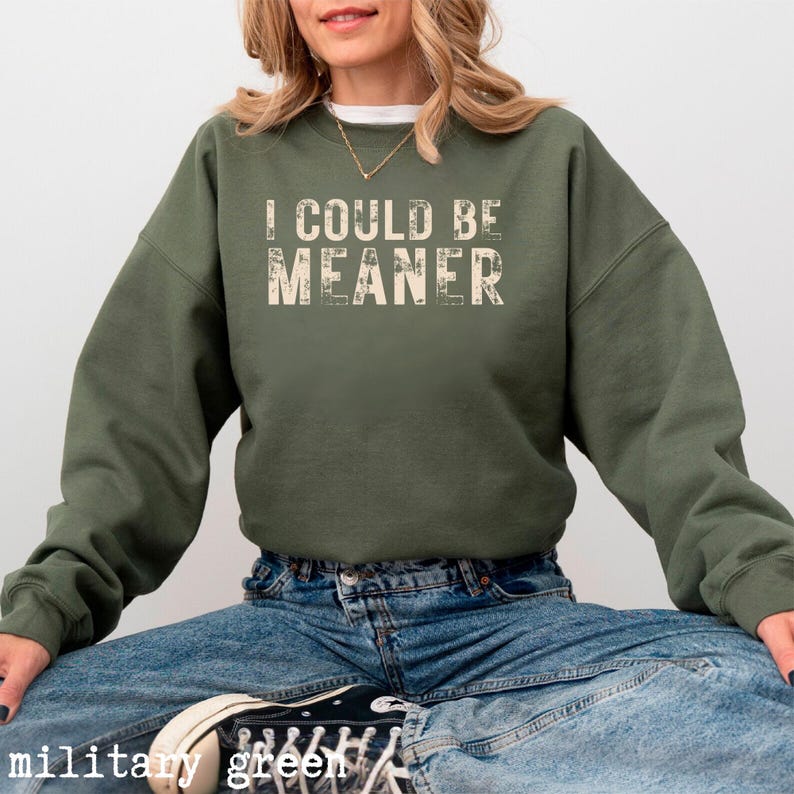 May include: Military green sweatshirt with the text "I COULD BE MEANER" in a distressed font. The sweatshirt is a solid color and has long sleeves. The model is wearing blue jeans and black sneakers.