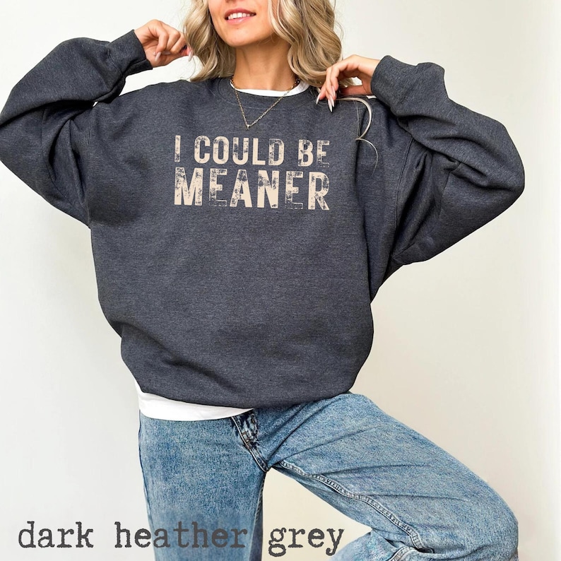 May include: Dark heather grey sweatshirt with the text "I COULD BE MEANER" in a distressed font. The sweatshirt is a relaxed fit and has long sleeves. The bottom of the image shows the words "dark heather grey".