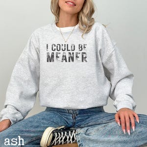 May include: A light gray sweatshirt with the text "I COULD BE MEANER" in a distressed black font. The model is wearing blue jeans and black and white sneakers. The word "ash" is printed at the bottom left.