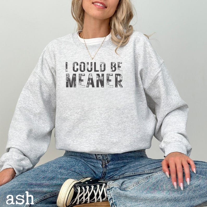 May include: A light gray sweatshirt with the text "I COULD BE MEANER" in a distressed black font. The model is wearing blue jeans and black and white sneakers. The word "ash" is printed at the bottom left.
