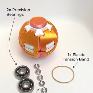 May include: A spherical orange and white object labeled INSIDE THE BALL, accompanied by its components: two silver precision bearings, five silver counterweight hex nuts, and one tan elastic tension band, all displayed with clear text labels.
