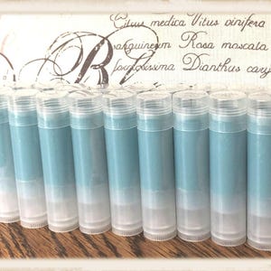 May include: A collection of blue lip balm tubes with clear caps. The tubes are arranged in a row on a wooden surface. The background features a decorative fabric with botanical text and a large letter "R".