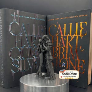 Può includere: Una scultura nera di una coppia che si abbraccia, posta davanti a due libri. I libri sono di Callie Hart, con titoli tra cui "Quick Silver" e "Brimstone". Il testo "#1 New York Times Bestseller" è visibile sulle copertine dei libri. Un adesivo recita "Made by a Book Lover".