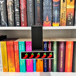 May include: A black metal shelf divider featuring a horizontal rectangular sign with five red chilli pepper icons with green stems arranged in a row against a black background, positioned between two rows of various hardcover books on white shelves.