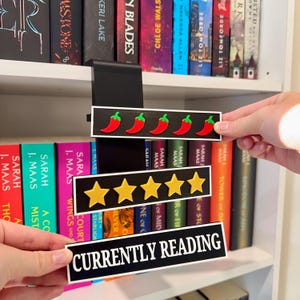 Puede incluir: Un marcador de estantería vertical negro con tres letreros rectangulares intercambiables. El letrero superior muestra cinco iconos de chiles rojos, el central cinco estrellas amarillas y el inferior el texto blanco CURRENTLY READING.