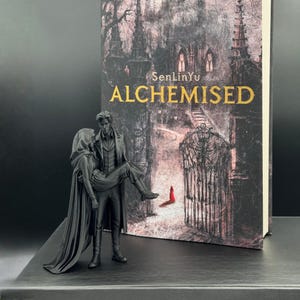 Könnte beinhalten: Eine schwarze Skulptur eines Mannes, der eine Frau trägt, vor einem Buch mit dem Titel "ALCHEMISED" von SenLinYu platziert. Das Buchcover zeigt eine gotische Illustration. Die Skulptur und das Buch stehen auf einer schwarzen Oberfläche.