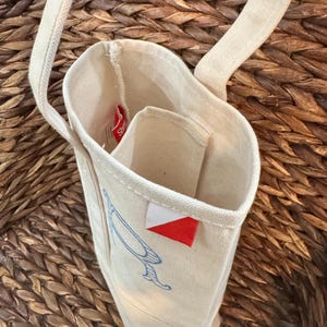 May include: A tall, off-white canvas wine tote bag with two handles and an open top. The exterior features a blue line-art graphic and a small, rectangular red and white fabric tag sewn onto the side seam. The interior includes a divider.