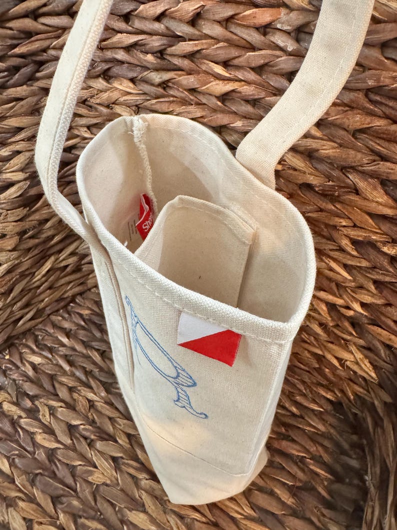 May include: A tall, off-white canvas wine tote bag with two handles and an open top. The exterior features a blue line-art graphic and a small, rectangular red and white fabric tag sewn onto the side seam. The interior includes a divider.