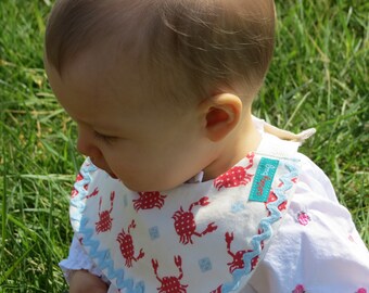 May include: A baby wearing a white bib with a blue scalloped edge and a red crab pattern. The bib has a blue label that says "baby bug".