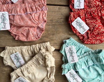 May include: Four pairs of baby bloomers with white floral patterns. The bloomers are in pink, beige, red, and light blue. Each pair has a white tag with the text "ashleybug originals" on it.
