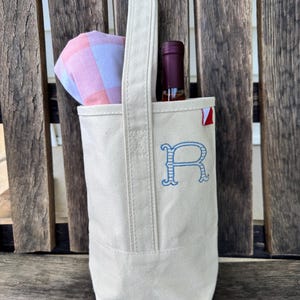 May include: A cream-colored canvas wine tote bag featuring a single tall handle and a blue embroidered letter R on the front. A wine bottle with a dark red cap and a folded pink and white checkered cloth are tucked inside the open top of the bag.