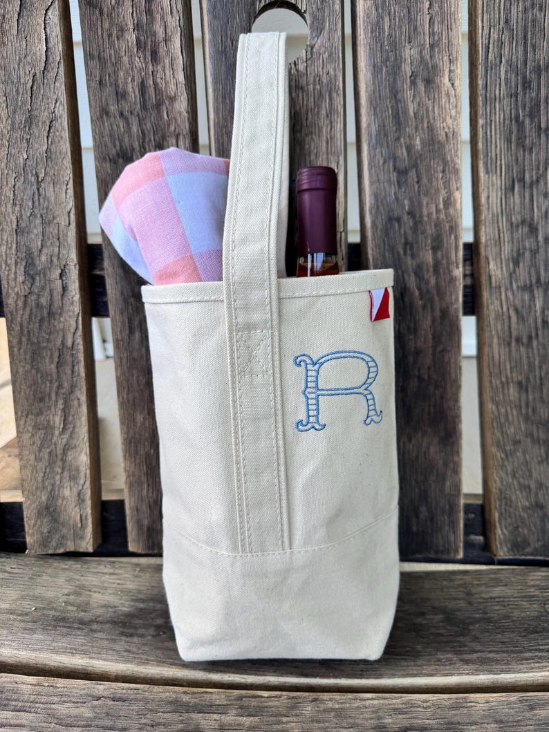 May include: A cream-colored canvas wine tote bag featuring a single tall handle and a blue embroidered letter R on the front. A wine bottle with a dark red cap and a folded pink and white checkered cloth are tucked inside the open top of the bag.
