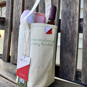 May include: A cream-colored canvas wine tote bag featuring a single handle and the embroidered green text "I never show up empty handed." A wine bottle and a pink and blue plaid cloth are inside. A ShoreBags brand tag with a QR code is attached.
