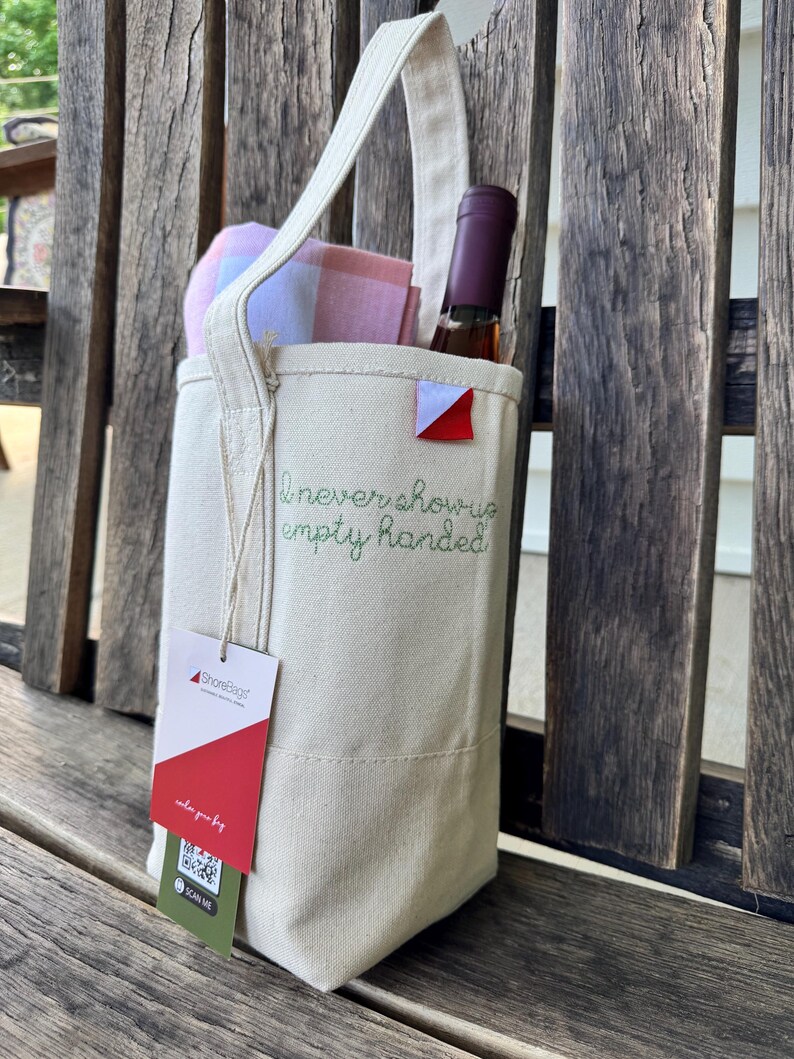 May include: A cream-colored canvas wine tote bag featuring a single handle and the embroidered green text "I never show up empty handed." A wine bottle and a pink and blue plaid cloth are inside. A ShoreBags brand tag with a QR code is attached.