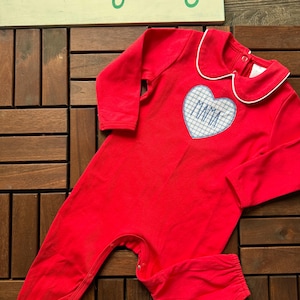 May include: Red baby romper with a white-trimmed collar and a heart-shaped patch. The patch features the word "MAMA" in blue embroidery. The romper has long sleeves and snaps for easy changes. The brand name "ashleybug" is visible.